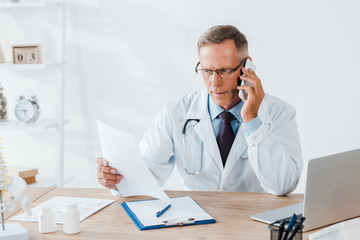 doctor in glasses and white coat talking on smartphone and looking at paper