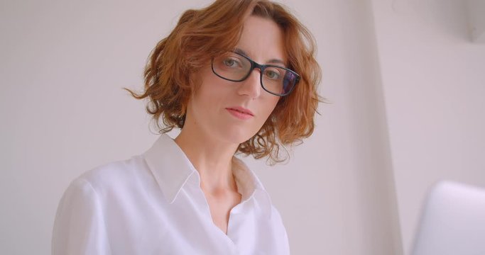 Closeup portrait of redhead caucasian businesswoman in glasses using laptop looking at camera smiling sitting in armchair in white apartment