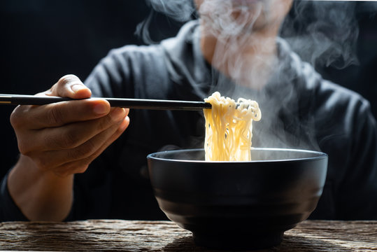 Hand uses chopsticks to tasty noodles with steam and smoke in bowl on wooden background, selective focus. Asian meal on a table, junk food concept