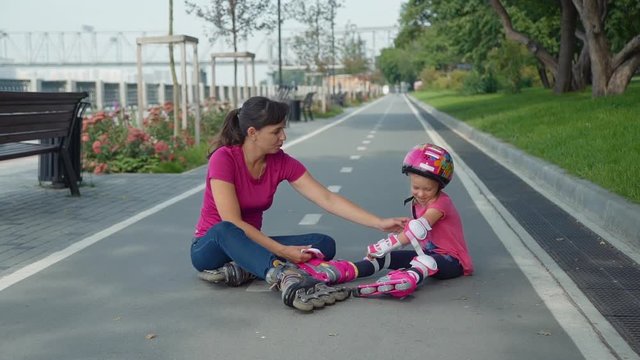 Mother and her Daughter Sitting on the Bicycle Lane after Rollerblading in a City Park. Little Girl in the Colorful Sport Helment Taking Off her Protective Gear with Help of her Mother.