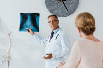 selective focus of doctor in white coat and glasses holding pen near x-ray and woman in clinic