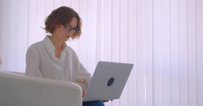Closeup side view portrait of adult redhead caucasian businesswoman in glasses using the laptop looking at camera sitting in the armchair in the white office