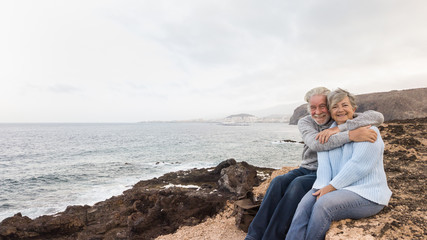 Fototapeta na wymiar Two senior people sitting on the cliff of the ocean and looking at camera Hugging and smiling. Morning soon outside, ready for un healthy excursion. Vacation and happiness