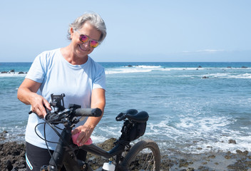 Senior woman grandmother with gray hair and the blue sea behind her cleaning her bicycle. Enjoying e bike activity for healthy lifestyle. Waves and rocks. Happy woman with sunglasses under the sun