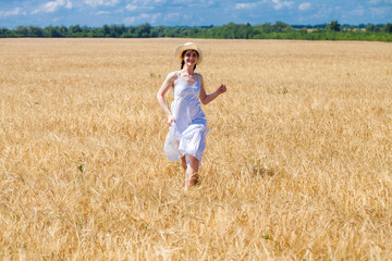 Naklejka na ściany i meble Happy brunette woman in white dress and straw hat walking in a wheat field