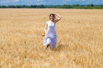 Naklejka na ściany i meble Happy brunette woman in white dress and straw hat walking in a wheat field