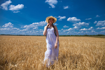 Happy brunette woman in white dress and straw hat walking in a wheat field