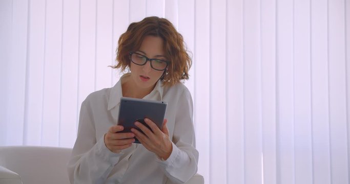 Closeup portrait of adult redhead caucasian businesswoman in glasses using the tablet sitting in the armchair in the white office