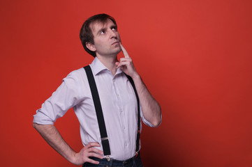 handsome confident man with hand on hip and finger on cheek in pink shirt and black suspender looking away and thinking on orange background with copy space