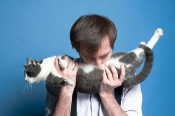 handsome man in shirt and black  suspender holding and kissing stomach cute gray and white cat on blue background