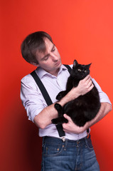 handsome man in shirt with rolled up sleeves and suspender holding black cat, smiling and looking at it on coral background with copy space