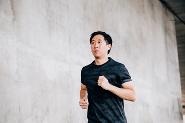 Serious Asian male athlete running on city street under grungy concrete bridge during outdoor training
