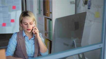 Tracking left of young blonde woman in office wear sitting at desktop, having phone conversation, writing note on sticker and pasting it on glass wall