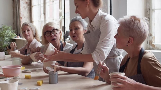 Tilt up of middle-aged female novice ceramists in aprons sitting at table, painting clay cups and watching young female tutor standing nearby showing them how to paint stoneware correctly