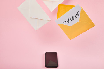 yellow envelope with white card with thanks lettering near letters and smartphone with blank screen on pink background
