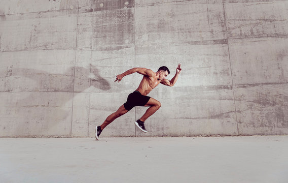Muscular shirtless caucasian male athlete does an explosive starting sprint against a grungy concrete wall background