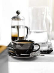 Coffee in black Cup on white background, table. French press,carafe of water, napkin, sunlight. Breakfast.