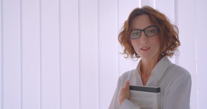 Closeup portrait of adult redhead caucasian businesswoman in glasses holding a book looking at camera smiling happily in the white apartment