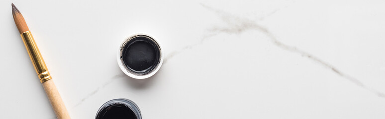 top view of black gouache paint and paintbrush on marble white surface with copy space, panoramic shot