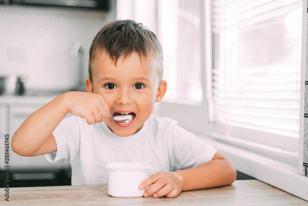 Wall mural cute boy in the kitchen eating yogurt from a white yogurt container, a place for advertising