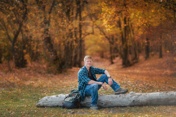 Redhead man in a plaid shirt and purple t-shirt sits on a tree log in autumn forest. Autumn, Nature, Outdoors, Recreation, Travel and Tourism concept.