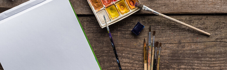 Obraz premium top view of colorful paint palette, paintbrushes and blank sketch pad on wooden surface, panoramic shot