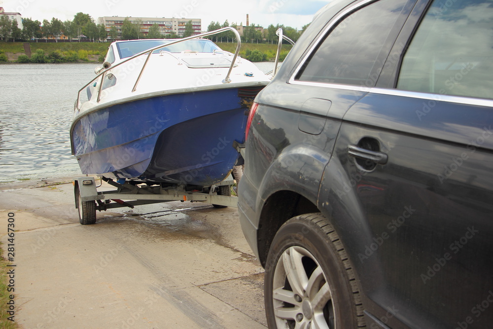 Wall mural A 4x4 black car pulls blue-white trimaran cabin motor boat on trailer on concrete slipway into the river water, DIY boat launch