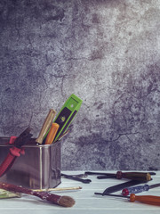 Various tools (screwdrivers, brush, wrench and others) are located in a metal box with rounded edges. Nearby are other tools. White wooden table. Stone or cement background with cracks.