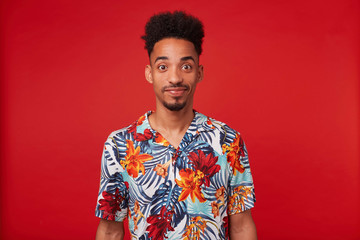 Young African American man wears in Hawaiian shirt, looks at the camera and calm smiling, stands...