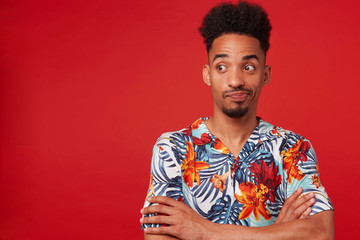 Young unhappy African American guy wears in Hawaiian shirt, looking left at the copy space, stands over red background with crossed arms.