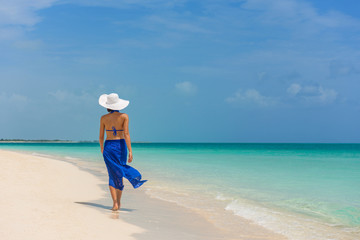 Luxury travel woman walking on perfect beach vacation Caribbean destination in blue dress skirt sarong. Elegant lady tourist relaxing on summer vacations wearing sun hat.