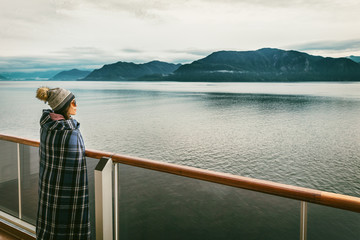 Alaska cruise travel luxury vacation woman watching inside passage scenic cruising day on balcony deck enjoying view of mountains and nature landscape. Asian girl tourist with wool blanket.