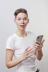 Fototapeta premium Happy funny millennial woman's celebrating win or victory, triumph, holding a phone. Cheerful excited girl, laughing, having fun, using cell phone apps. Isolated on white studio blank background.