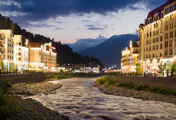 Fototapeta premium Panoramic view of Mzymta river and hotels and buildings at mountains background in Rosa Khutor resort in Krasnaya polyana, Sochi, Russia sunset time