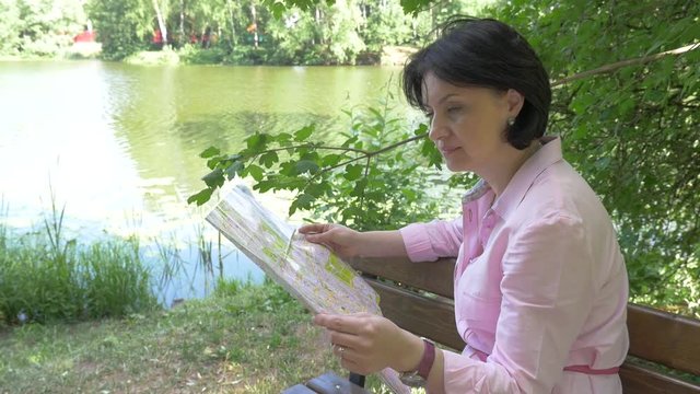 A middle-aged woman, a brunette in a pink shirt and sunglasses, sits on a park bench near a pond and looks at a paper map of tourist attractions. On a sunny summer day.