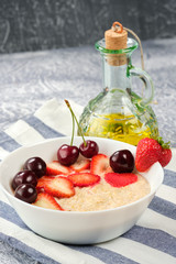 white bowl with oatmeal and strawberries and cherries and olive oil on a striped napkin and a gray background.