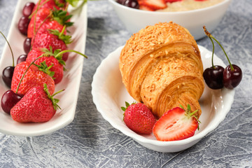 white bowl of porridge with strawberries and cherries, croissants and crispbread with cheese and strawberries on a gray background