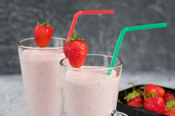 Two glasses with strawberry milkshake with pieces of berries on the edge of the glass on gray background.