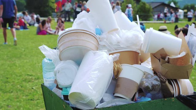 Plastic and paper trash in a crowded container in the park next to the cafe. In the background, people relax on the green lawn at a festival or village fair.