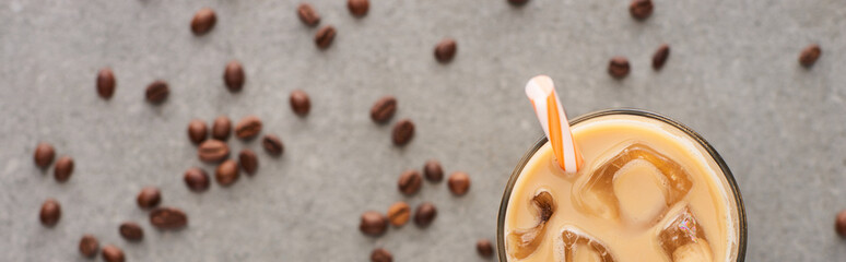 selective focus of ice coffee in glass with straw and coffee grains on grey background, panoramic shot