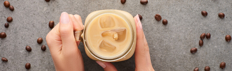 cropped view of woman holding glass jar with ice coffee and coffee grains on grey background, panoramic shot