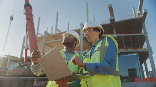Two Specialists Inspect Commercial, Industrial Building Construction Site. Real Estate Project with Civil Engineer, Investor Use Laptop. In the Background Crane, Skyscraper Concrete Formwork Frames 
