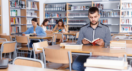 Portrait of young bearded man sitting in public library and reading books