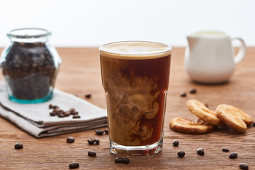 selective focus of ice coffee mixing with milk in glass near milk jug, cookies and coffee grains on...