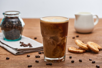 selective focus of ice coffee mixing with milk in glass near milk jug, cookies and coffee grains on wooden table isolated on grey