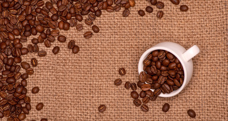 Coffee grains are scattered on a jute napkin.  Nearby is an overturned cup of coffee beans.Texture background. Copy space. The concept of cheerfulness and good mood.