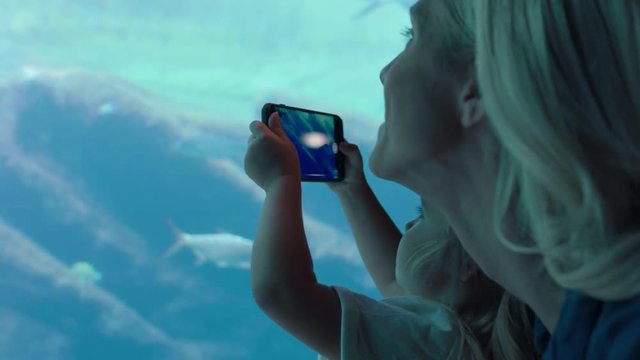 girl taking photo of fish in aquarium using smartphone mother and daughter photographing marine life swimming in tank with mobile phone camera having fun at oceanarium sharing on social media