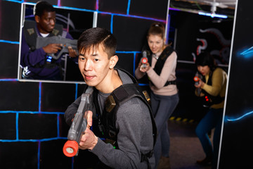 Emotional Asian man with laser pistol playing laser tag with friends on dark labyrinth