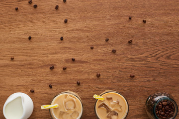 top view of coffee in glasses with ice and straws, milk and coffee grains on wooden table with copy space