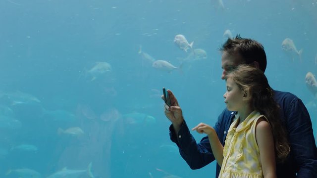father with little girl at aquarium taking photos of fish using smartphone enjoying marine life swimming in tank having fun at oceanarium sharing on social media